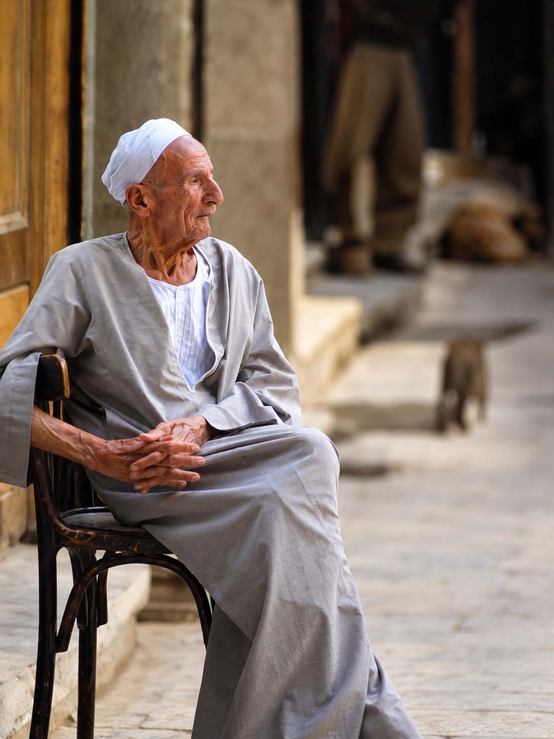 رجل مصري مسن يرتدي الجلباب التقليدي والعمامة البيضاء يجلس على كرسي خشبي في ممر تاريخي بشارع المعز — صورة 2